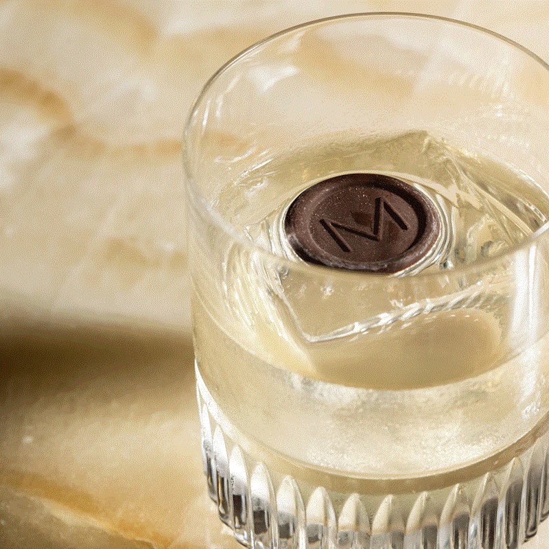Clear cocktail in a rocks glass with a chocolate coin garnish stamped with an M