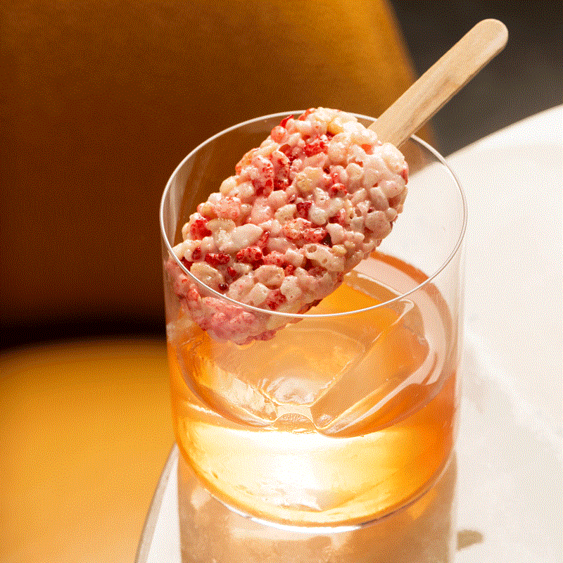 Cocktail in a rocks glass with red and pink marshmallow crispy rice treat on a stick as the garnish