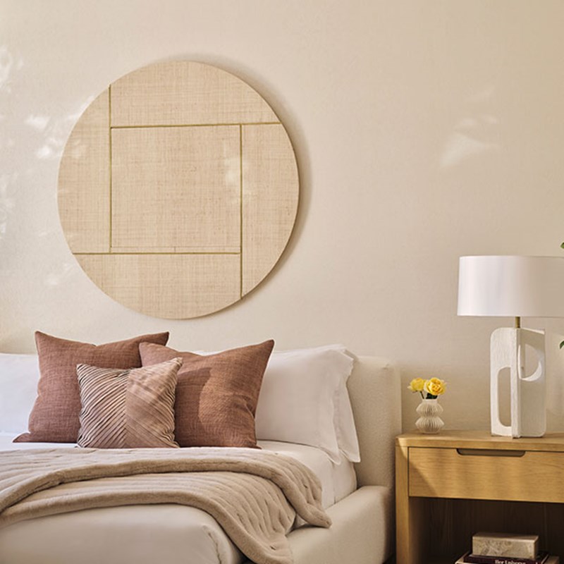 Corner of a double bed with earthy tone bedding and cushions, and a table and light beside it