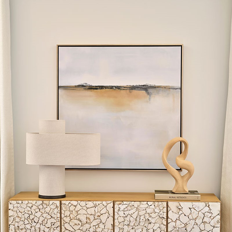 Sideboard with a neutral colour piece of art behind it and a lamp and sculpture on it